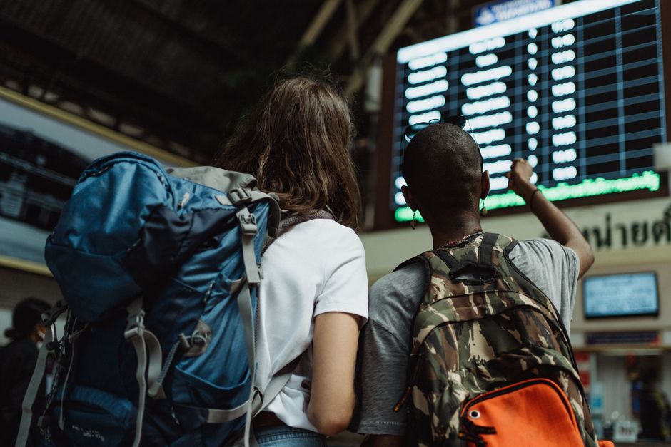 A solo traveler, wearing a backpack, standing on a train platform in Europe, looking at a departure board and checking a mobile phone, representing flexible travel.