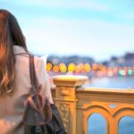 A solo female traveler smiles while browsing fresh produce at a vibrant European market, her small backpack beside her.