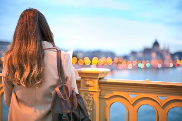 A solo female traveler smiles while browsing fresh produce at a vibrant European market, her small backpack beside her.