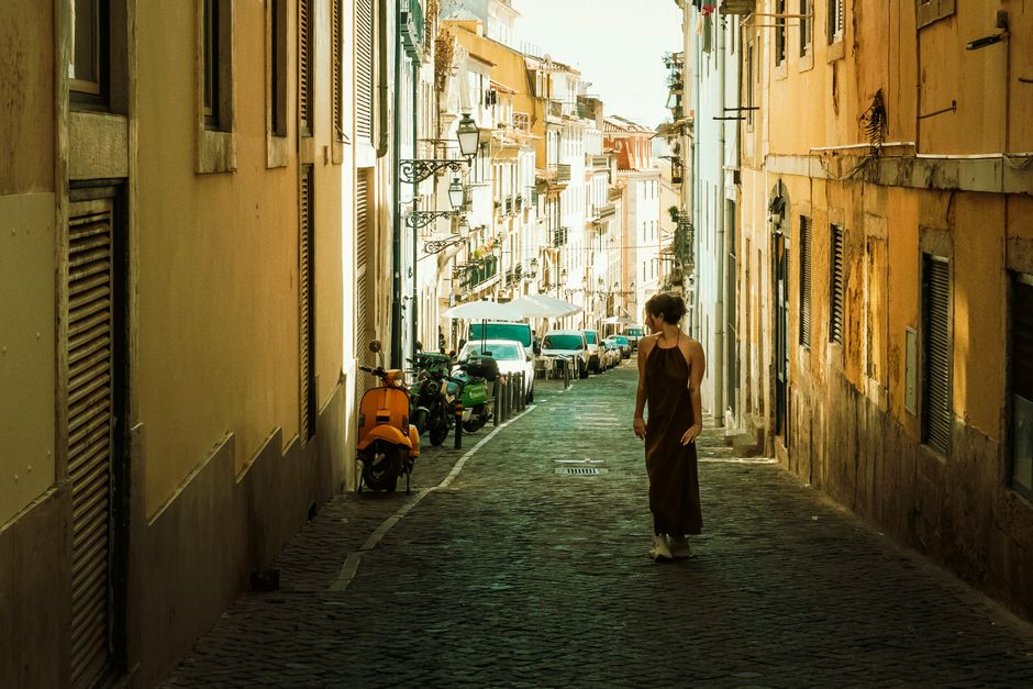 A solo female traveler from North America, looking at a map while walking through a charming, narrow cobblestone alley in Lisbon, Portugal, illustrating cultural immersion.