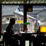A solo woman traveler sips coffee peacefully at an outdoor cafe, with a travel journal and phone on the table, indicating a relaxed morning.