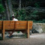 A solo traveler reads a book on a park bench, surrounded by green trees and sunshine, embodying slow travel.