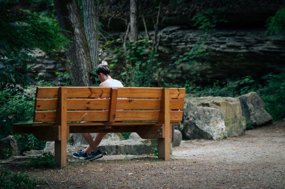 A solo traveler reads a book on a park bench, surrounded by green trees and sunshine, embodying slow travel.