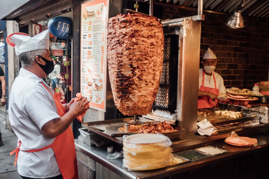 A street food vendor in Mexico City expertly grilling and assembling tacos on a bustling street, with steam rising and colorful garnishes visible.