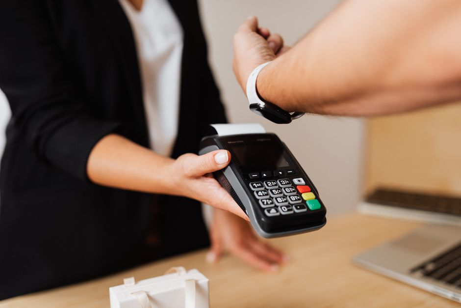 A student paying for a coffee using their smartwatch at a bustling cafe counter, card machine visible.