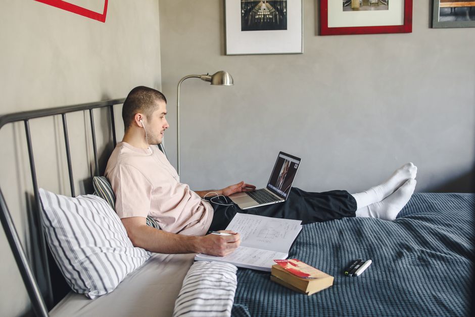 A student studying or working remotely on a laptop in a cozy, confined living space, possibly a bedroom corner, with books and a coffee mug nearby.