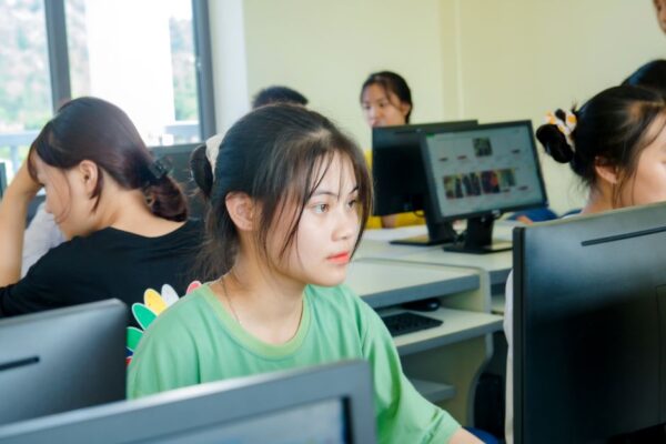 A young woman, likely a student, focused on a modern monitor while taking notes in a well-lit study space with books and a laptop.