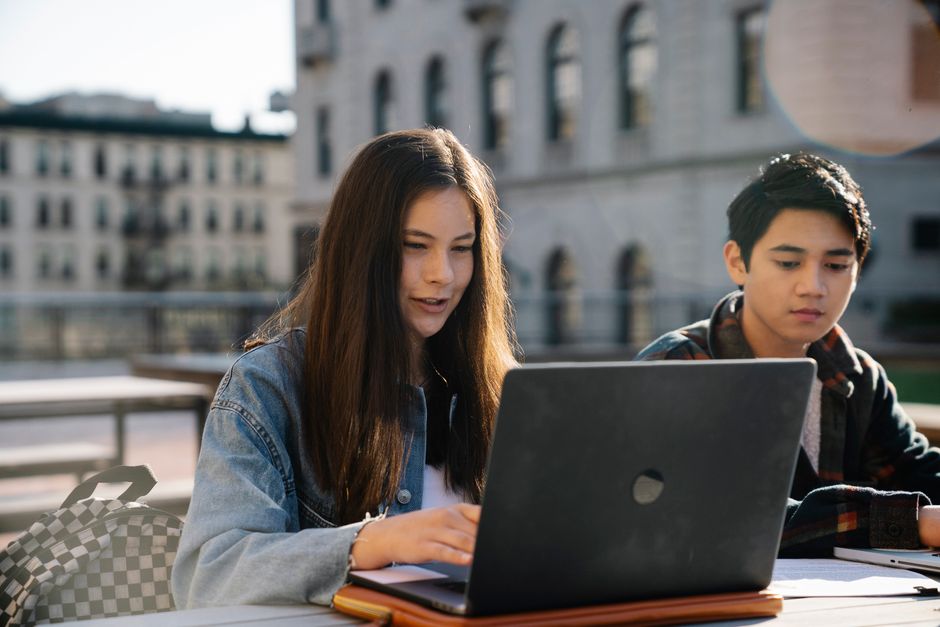 A student with glasses focused on a laptop screen, showing an AI chat interface, surrounded by open textbooks and notes.