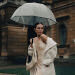 A stylish woman in a long, dark waterproof trench coat walking confidently down a city street in the rain, holding an umbrella, demonstrating effective rainy day style for commutes.