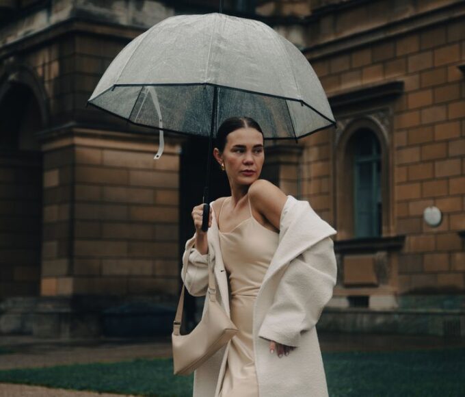 A stylish woman in a long, dark waterproof trench coat walking confidently down a city street in the rain, holding an umbrella, demonstrating effective rainy day style for commutes.