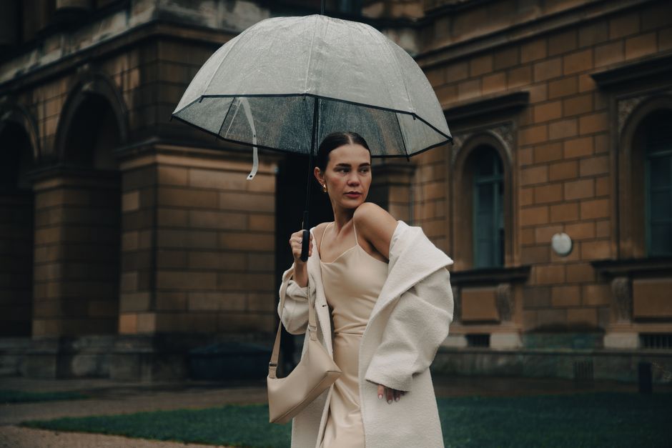 A stylish woman in a long, dark waterproof trench coat walking confidently down a city street in the rain, holding an umbrella, demonstrating effective rainy day style for commutes.