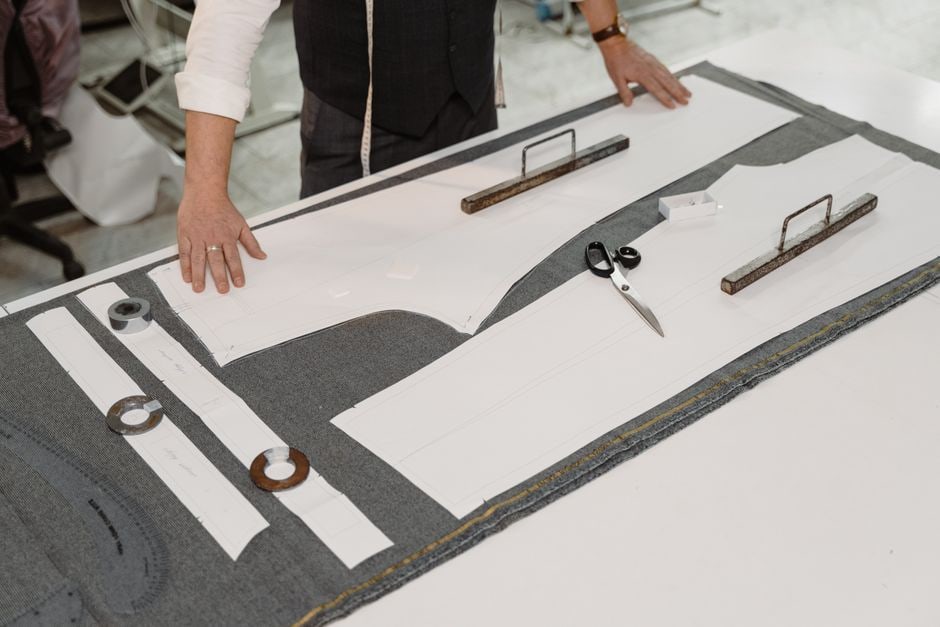 Close-up of a tailor's hands precisely stitching a seam on a pair of trousers, with a measuring tape visible.