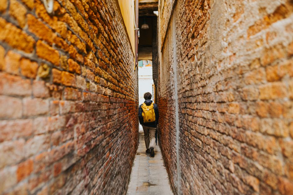 A person with a backpack and comfortable walking shoes explores a narrow, historic alleyway in a European city, looking up at old buildings, depicting the blend of comfort and style needed for city travel.