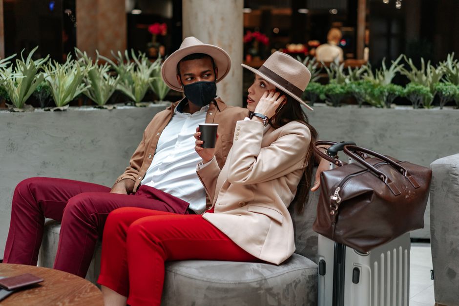 A person in an airport lounge, dressed in a comfortable yet chic neutral outfit, including a cashmere travel wrap and simple gold jewelry.