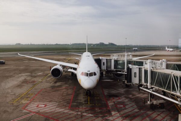 A traveler in comfortable clothes doing a standing leg stretch near a large airport window, with planes visible outside.