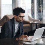 A remote worker sitting in a bustling European cafe, surrounded by travel tech, actively working on a laptop with a smartphone charging nearby.