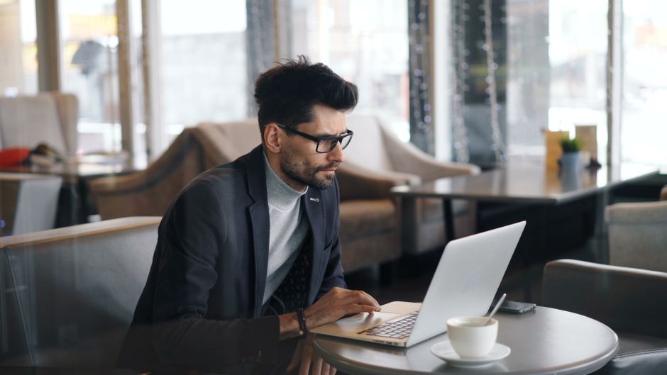 A remote worker sitting in a bustling European cafe, surrounded by travel tech, actively working on a laptop with a smartphone charging nearby.