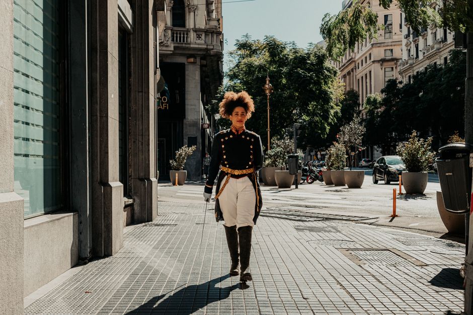A stylish professional woman confidently walking down a bustling European city street, wearing a smart blazer, tailored trousers, and comfortable sneakers.