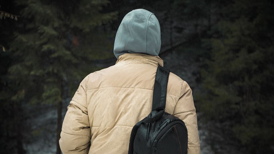 A traveler wearing a sleek, modern, olive green travel jacket with subtle features, standing in an outdoor setting like a park or urban plaza, demonstrating its versatility.