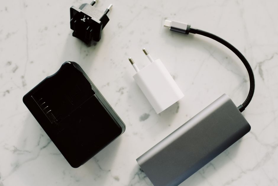 A hand holding a USB-C cable connecting to a laptop, with other USB-C devices like a phone and tablet in the background.
