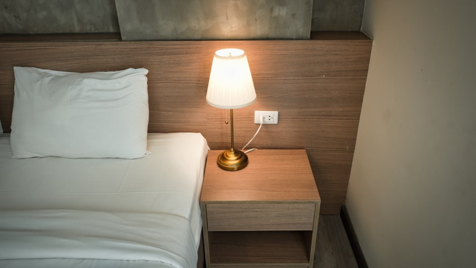 A compact white noise machine sits on a hotel room nightstand next to a lamp, casting a soft glow in a dimly lit room.