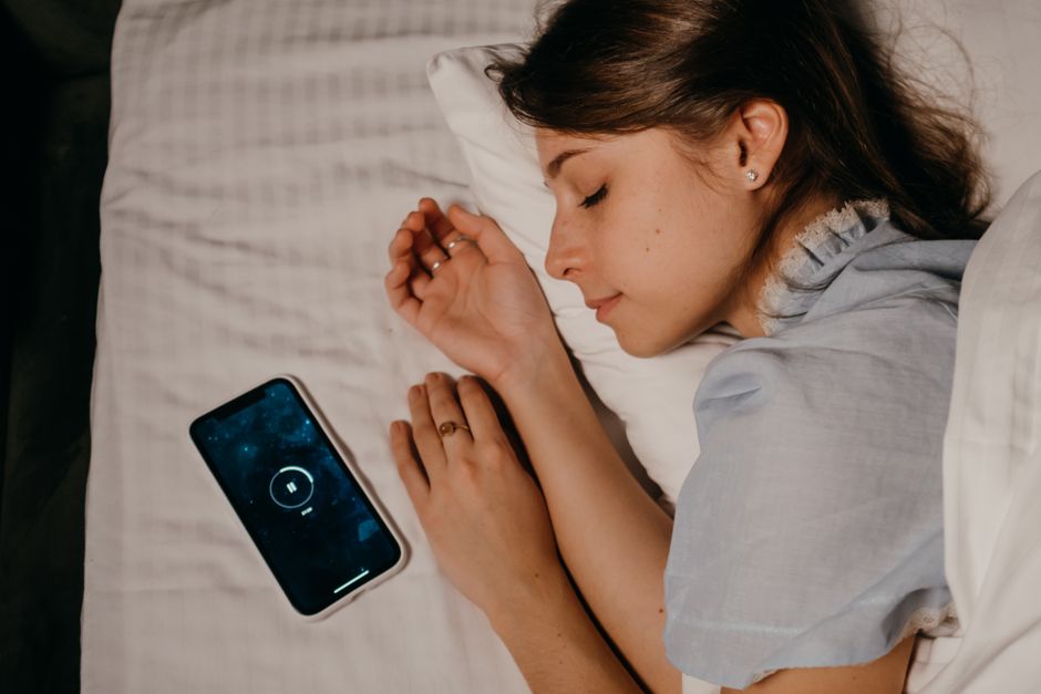 A woman in bed checking her sleep data on a smartphone, with an Oura Ring visible on her finger.