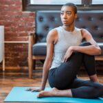 A woman in athletic wear performing a gentle yoga pose on a mat in a small, tidy living room with minimal furniture, emphasizing quiet movement.