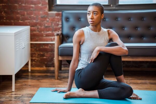 A woman in athletic wear performing a gentle yoga pose on a mat in a small, tidy living room with minimal furniture, emphasizing quiet movement.