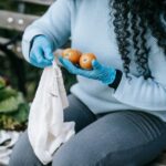 A busy woman smiling, holding a reusable canvas bag full of fresh produce, shopping at an outdoor farmers market.