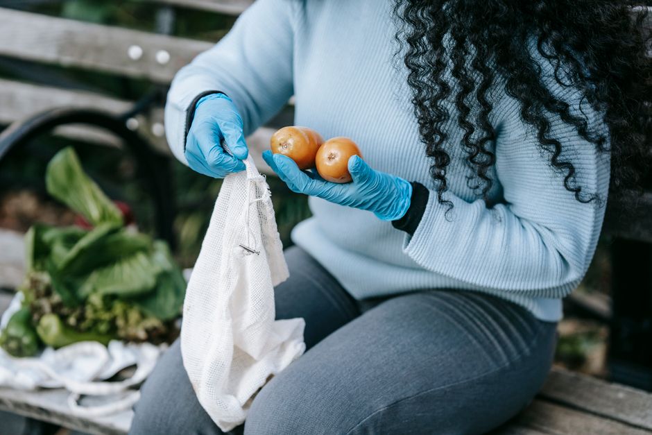 A busy woman smiling, holding a reusable canvas bag full of fresh produce, shopping at an outdoor farmers market.