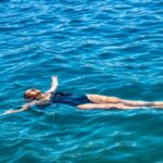 Woman wearing a stylish, sustainable one-piece swimsuit, looking out at the calm, blue sea, embodying relaxed summer travel.