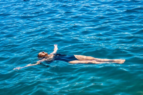 Woman wearing a stylish, sustainable one-piece swimsuit, looking out at the calm, blue sea, embodying relaxed summer travel.