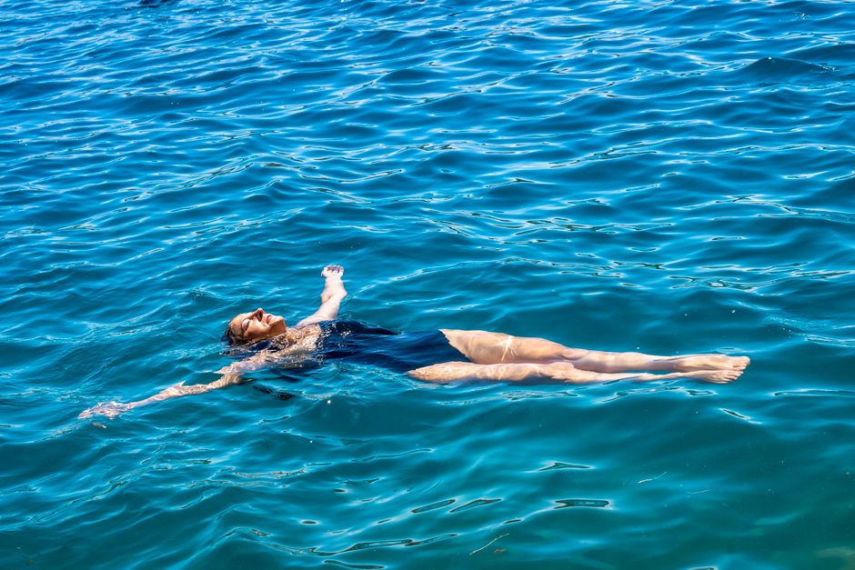 Woman wearing a stylish, sustainable one-piece swimsuit, looking out at the calm, blue sea, embodying relaxed summer travel.