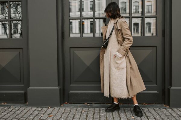 A stylish woman in London wearing a cream knit sweater, tailored camel trousers, and a dark long coat, demonstrating texture and neutral layering.