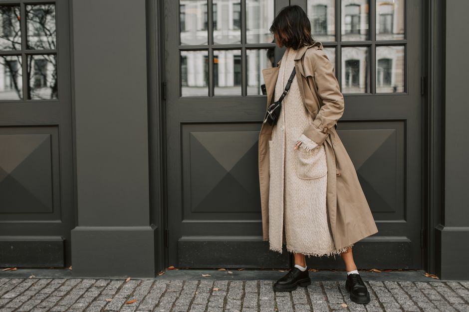 A stylish woman in London wearing a cream knit sweater, tailored camel trousers, and a dark long coat, demonstrating texture and neutral layering.