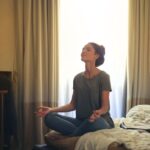A woman sits cross-legged on a rug in a softly lit bedroom, meditating peacefully as the sun sets outside the window, illustrating calm after screen-heavy days.