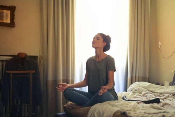 A woman sits cross-legged on a rug in a softly lit bedroom, meditating peacefully as the sun sets outside the window, illustrating calm after screen-heavy days.