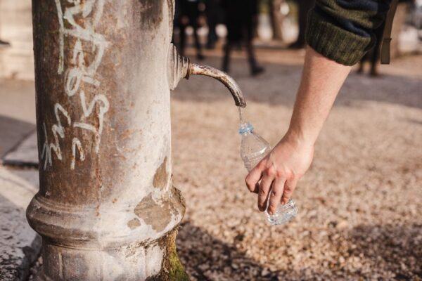A woman smiling as she refills her sleek, reusable water bottle at a modern public water fountain in a sunny city square.