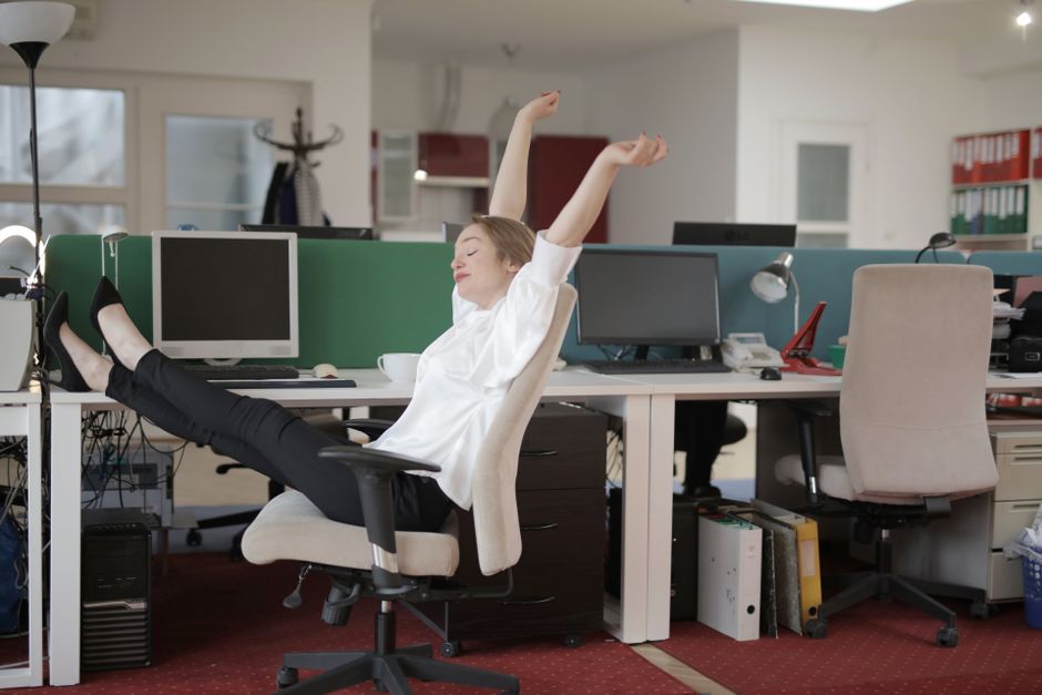 A woman in a modern office, wearing business casual attire, gently stretching her neck and shoulders while seated at her desk, with a laptop and plants visible.