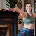 A woman in athletic wear performing a gentle stretch on a yoga mat in her bright living room, symbolizing home fitness.