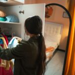 A woman with dark hair stands in front of an open, color-coded closet, thoughtfully touching a blouse. The closet is well-organized, reflecting a curated wardrobe.