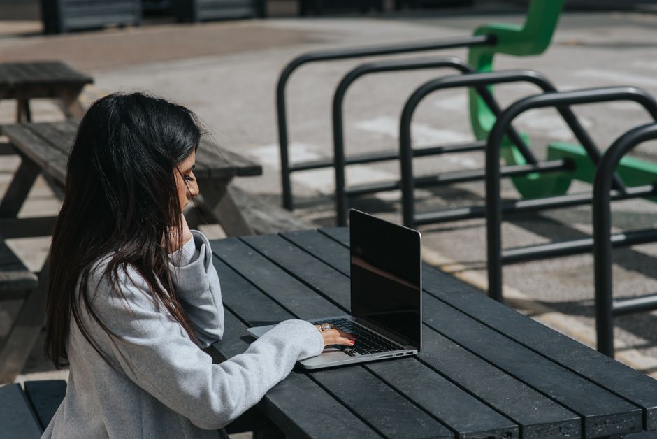 A woman smiling and working on her laptop while sitting outdoors in a bustling European city square.