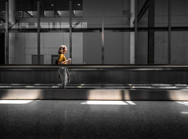 Woman in comfortable layers walking through a modern, brightly lit airport terminal, carrying a small travel bag and looking relaxed.