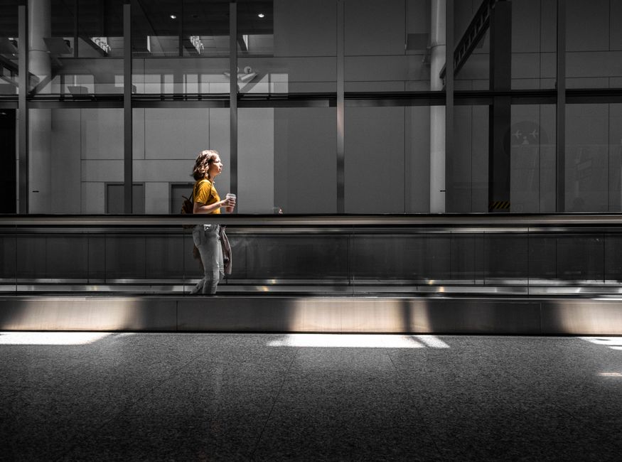 Woman in comfortable layers walking through a modern, brightly lit airport terminal, carrying a small travel bag and looking relaxed.