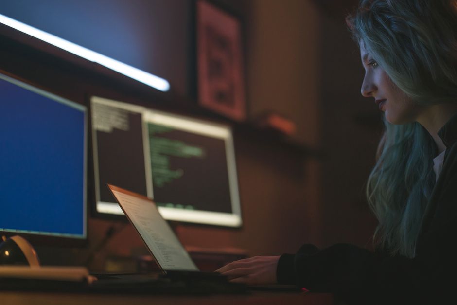 A woman sits at a desk in a dark room, illuminated by multiple computer screens, working late, illustrating the challenge of managing sleep hygiene with global work schedules.
