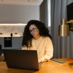 A woman working on a laptop at a small dining table in a cozy, bright apartment, illustrating the reality of small remote workspaces.