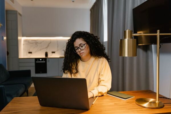 A woman working on a laptop at a small dining table in a cozy, bright apartment, illustrating the reality of small remote workspaces.