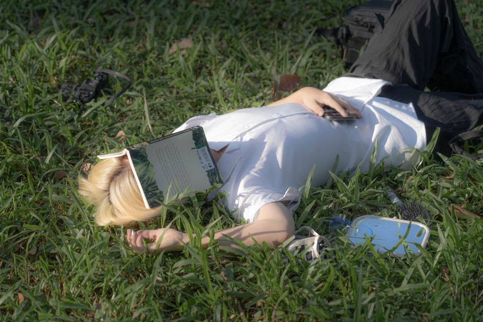 A student sitting on a park bench, reading a physical book and smiling, with their phone placed face down beside them, enjoying nature.